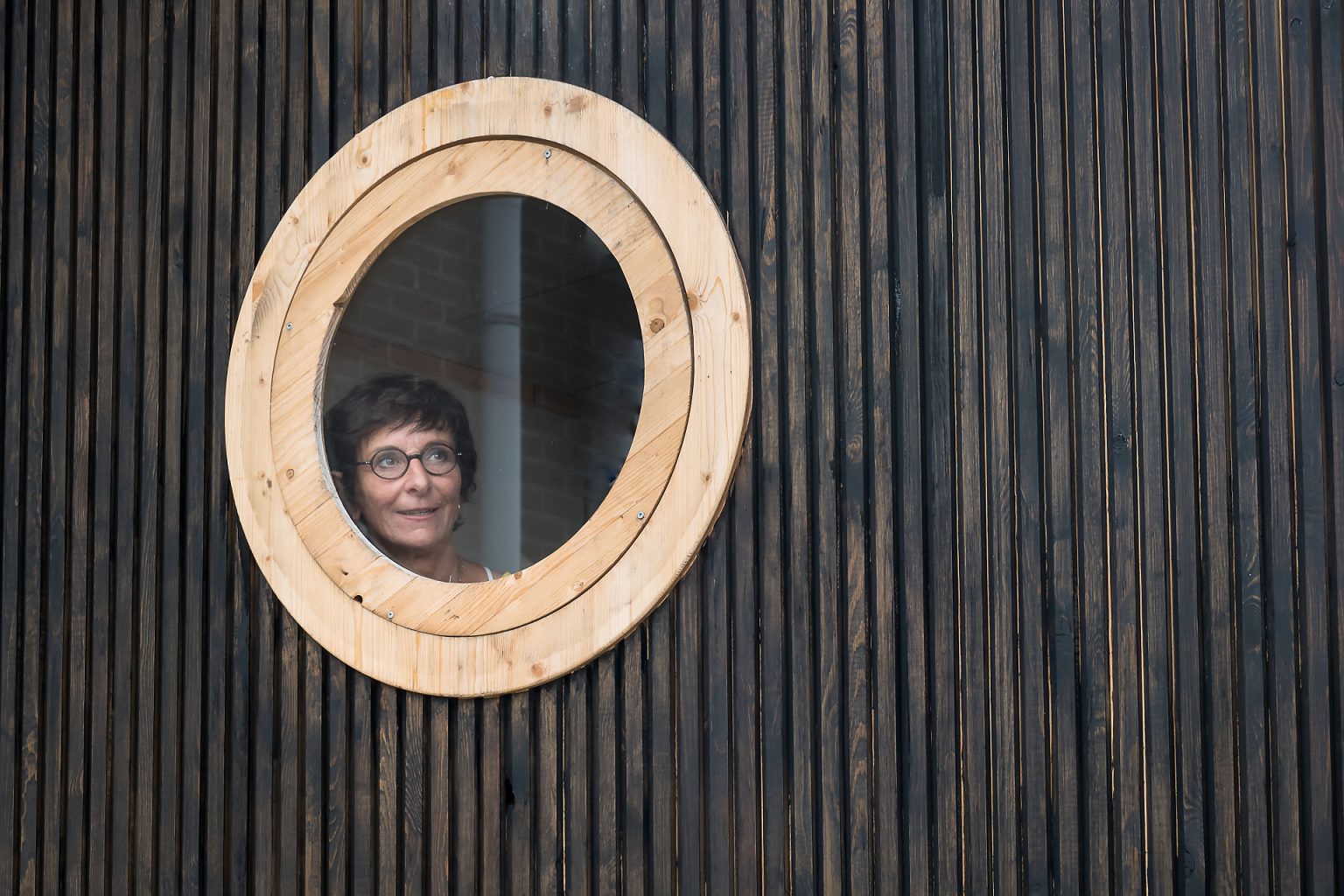 Portrait d'entrepreneur : Isabelle Dubois, architecte d'intérieur au hublot de sa roulotte en construction. Portrait en lumière naturelle et au plus proche de l'activité de l'entrepreneur