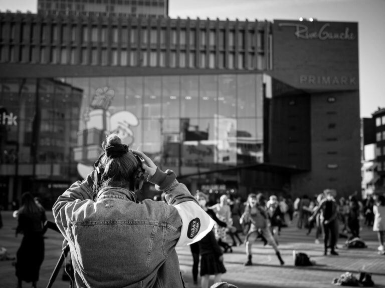 retrospective-2021 Une personne de dos avec des écouteurs face à une place animée en ville avec des passants et des bâtiments modernes