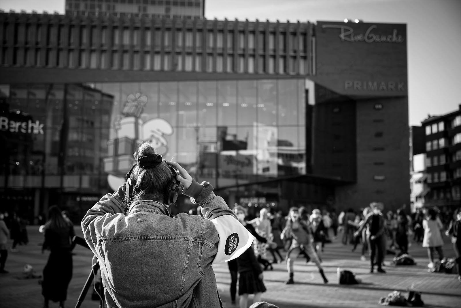retrospective-2021 Une personne de dos avec des écouteurs face à une place animée en ville avec des passants et des bâtiments modernes