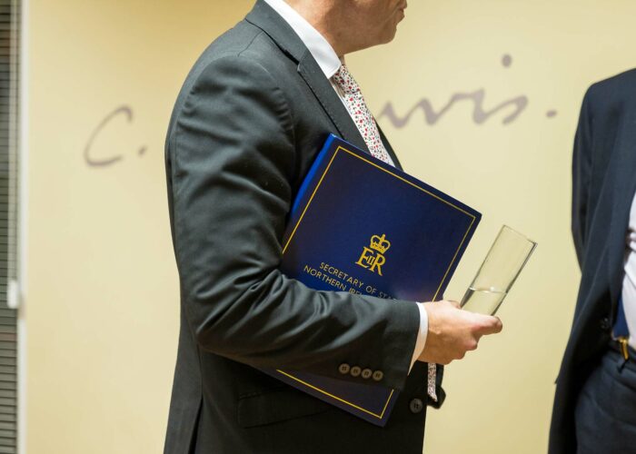 Un homme en costume sombre est vu de profil, tenant fermement un dossier bleu orné du logo royal "EIR" et de l'inscription "SECRETARY OF STATE NORTHERN IRELAND".