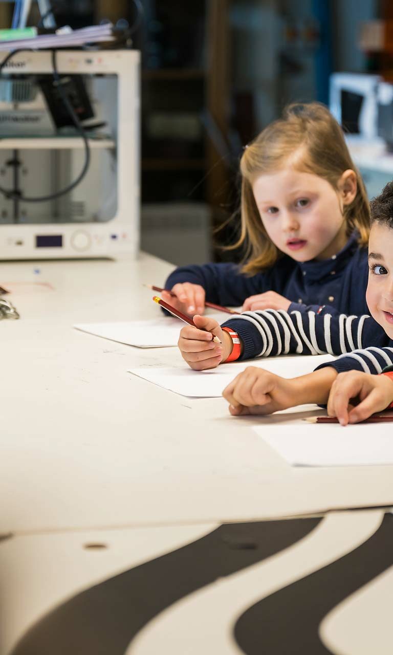 Un petit garçon souriant, un crayon à la main qui regarde l'objectif et deux petites filles qui sont à ses côtés