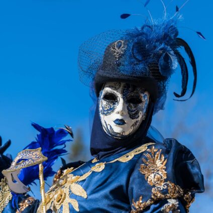 Une personne avec un costume bleu foncé avec des ornements dorés et des détails en dentelle et motifs brodés. La personne porte un masque vénitien blanc avec des motifs bleu et or