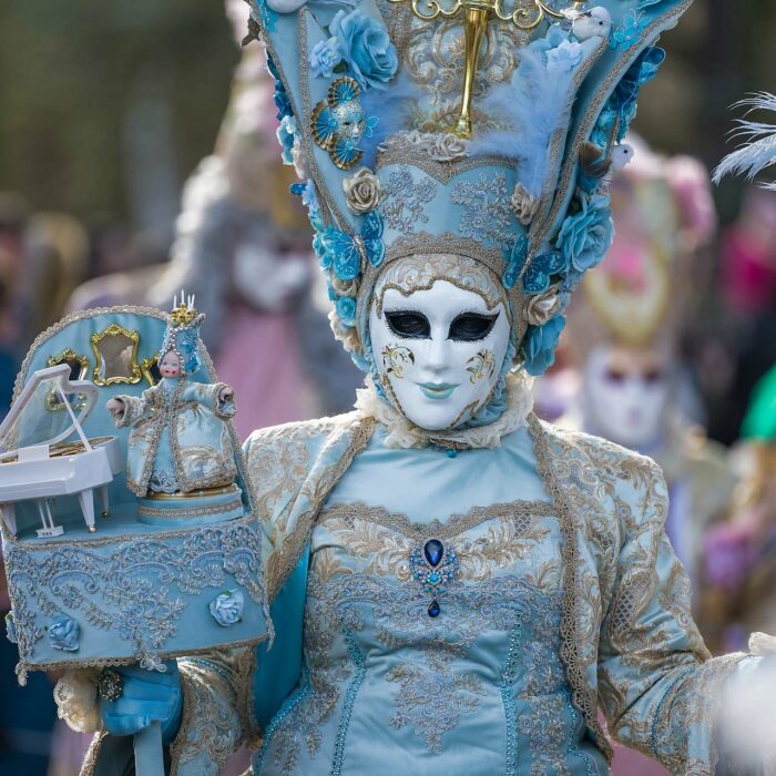 Une personne avec un costume élaboré bleu clair avec des ornements dorés et des détails en dentelle et motifs brodés. La personne porte un masque vénitien blanc avec des traits noirs au-dessus des yeux