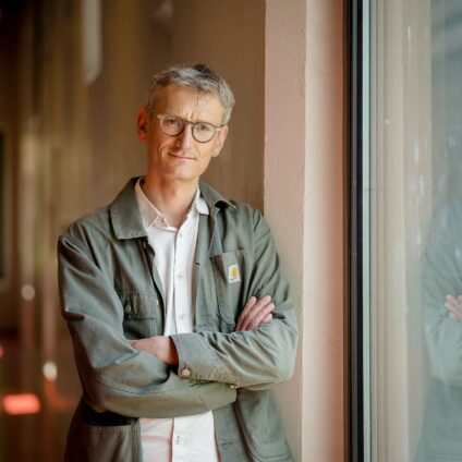 Portrait d'entreprise - Un homme avec des cheveux gris, des lunettes, une veste kaki qui se tient à côté d'une fenêtre