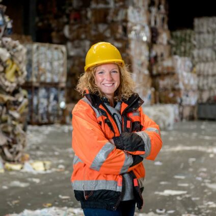 Portrait d'une ouvrière souriante vêtue d'un équipement de sécurité, incluant un casque de construction jaune et un gilet orange dans un centre de recyclage