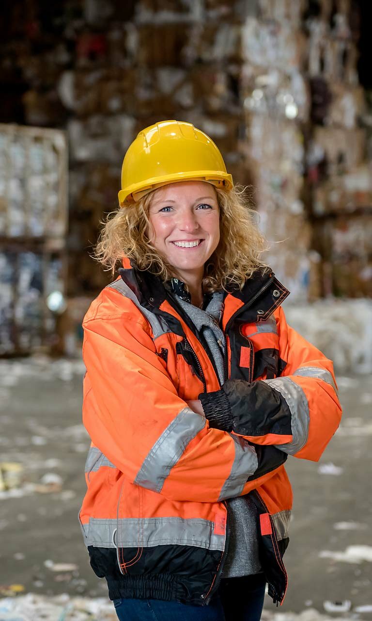 Portrait d'une ouvrière souriante vêtue d'un équipement de sécurité, incluant un casque de construction jaune et un gilet orange dans un centre de recyclage