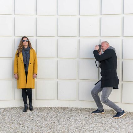 Une femme avec un manteau jaune qui se tient devant un mur aux carreaux blancs et un homme à sa droite qui capture son portrait