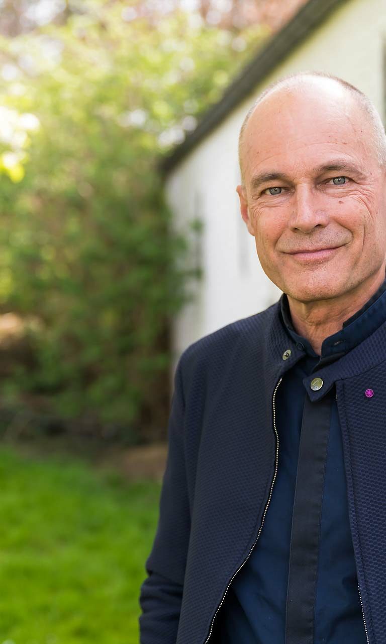 Portrait d'un homme qui pose devant un mur blanc et qui arbore une tenue élégante dans une veste bleu foncé