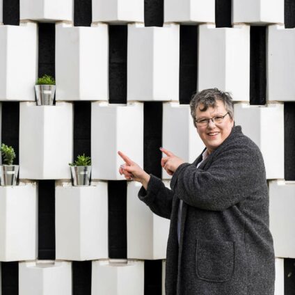 Une femme avec des lunettes et un long manteau gris foncé qui montre du doigt un mur avec des plantes en pots