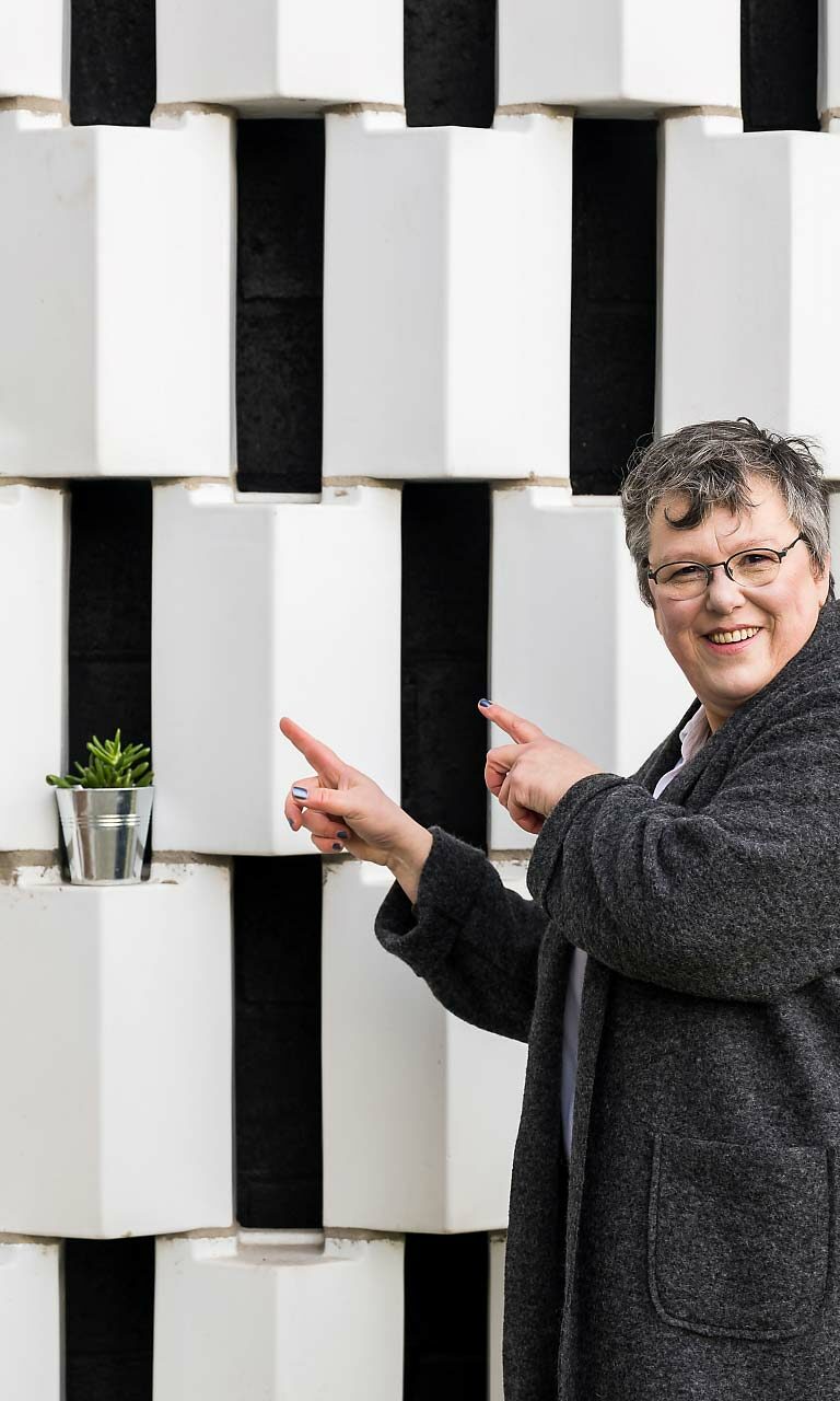 Une femme avec des lunettes et un long manteau gris foncé qui montre du doigt un mur avec des plantes en pots